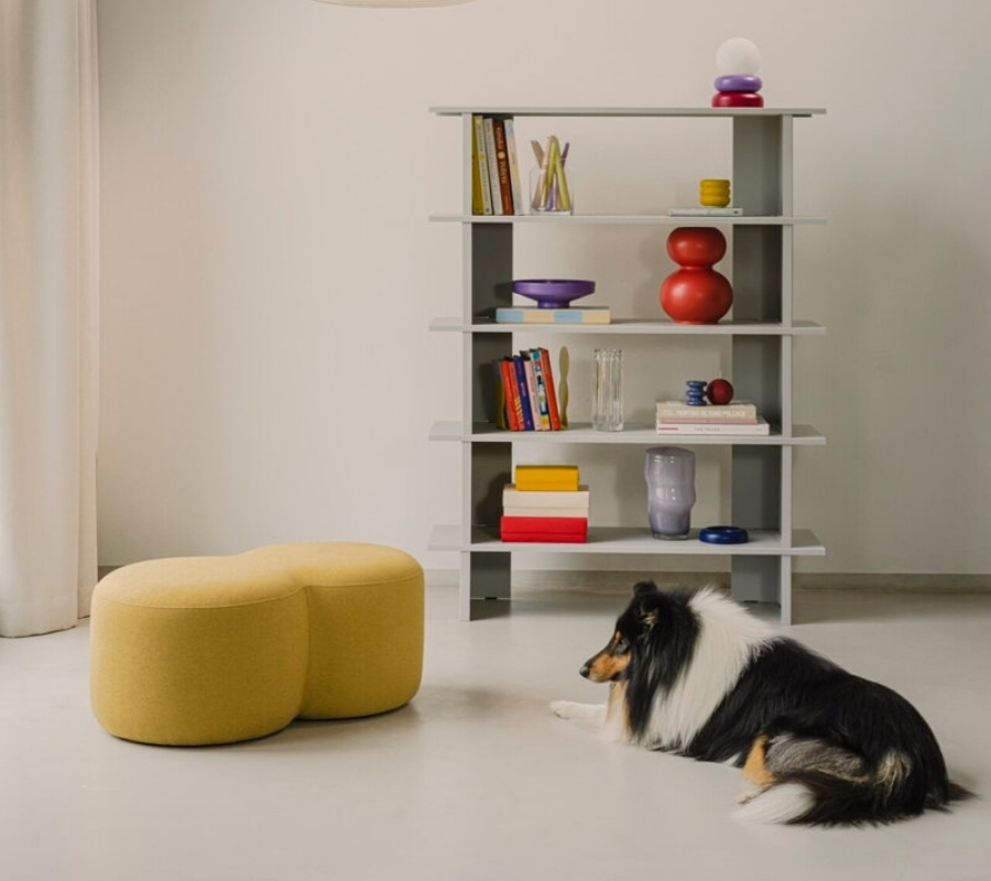Čalúnený zaoblený puf žltej farby pri oddychujúcom psovi a sivej knižnici s farebnými doplnkami.