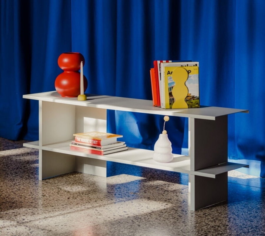 Nízka sivá polica z ocele s doplnkami a knihami, pri modrom závese.