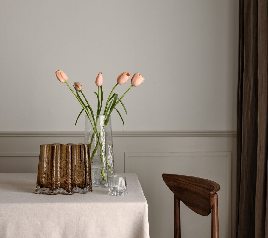 Číra váza z ručne fúkaného skla s kvetmi na stole pri hnedom svietniku.