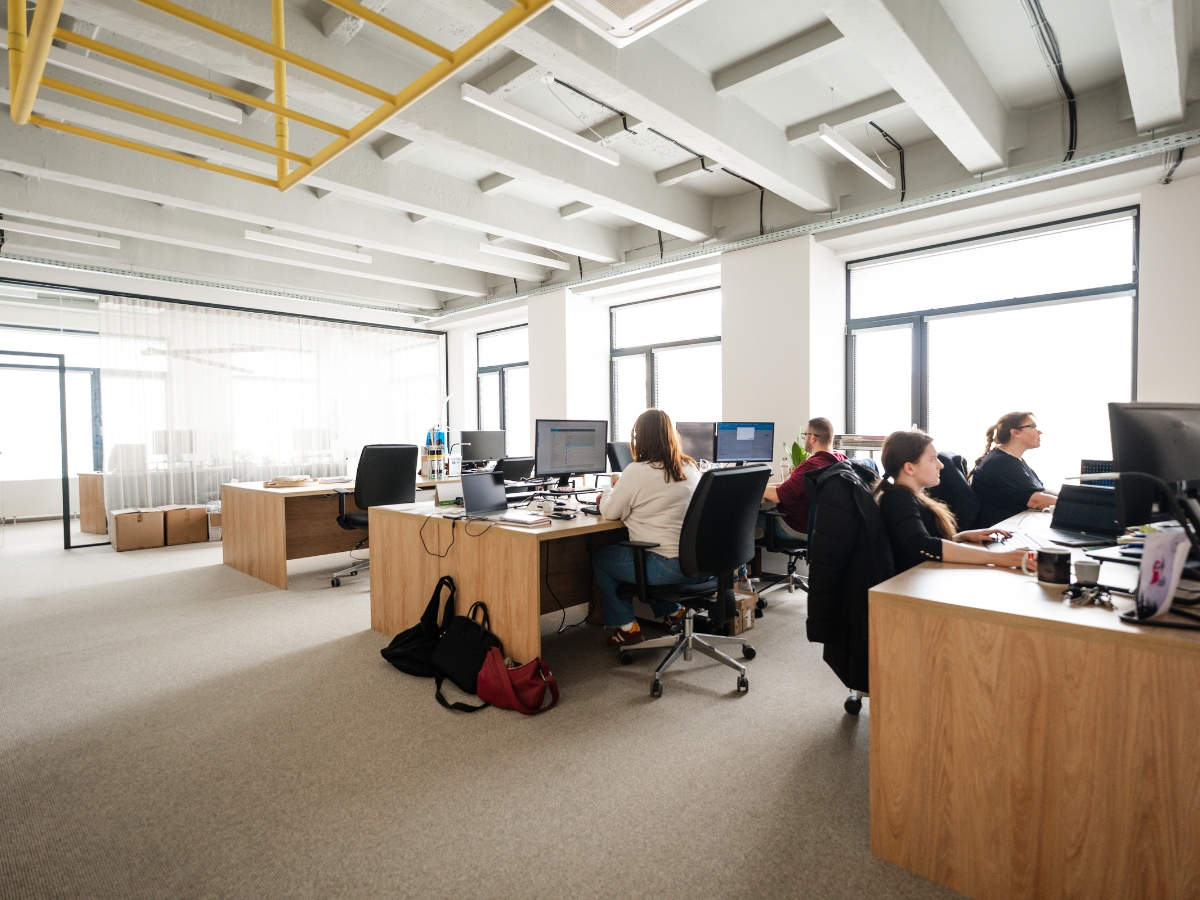 Open space office s viacerými pracovnými stolmi, veľkými oknami a štyrmi pracujúcimi ľuďmi.