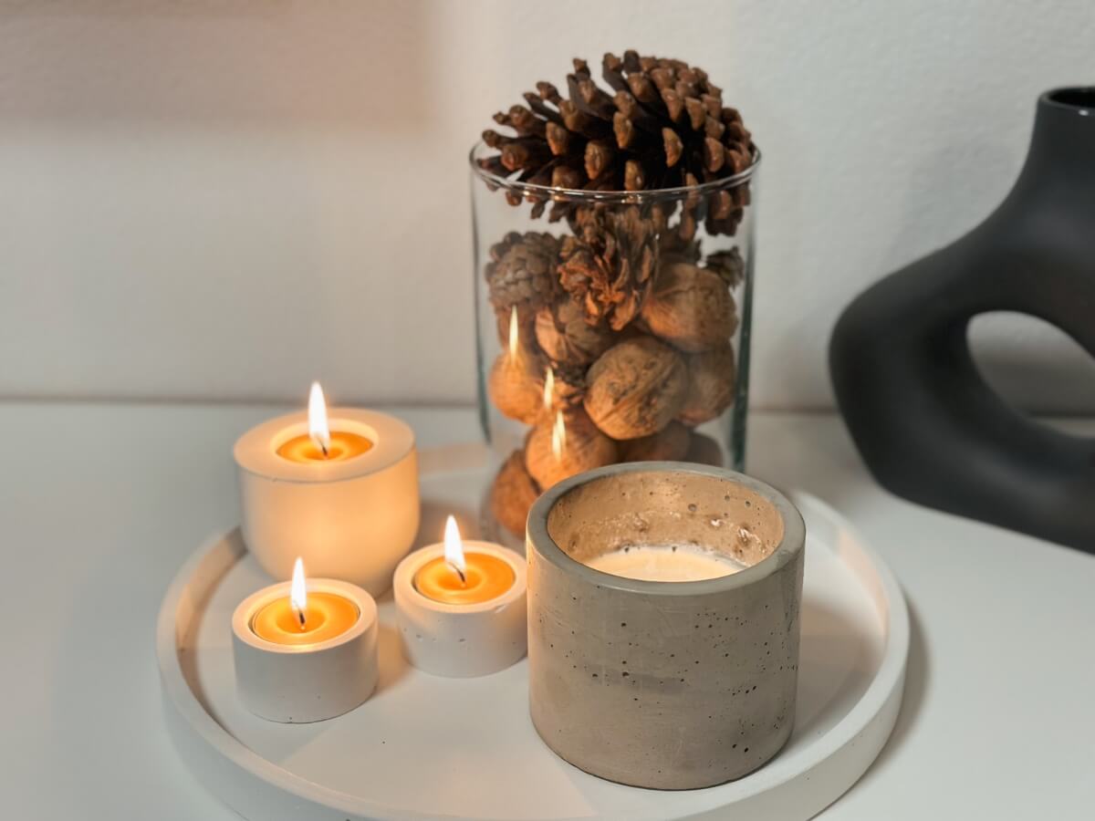 Horiace sviečky položené vedľa sklenenej nádoby s jesennou výzdobou šišiek a orechov.