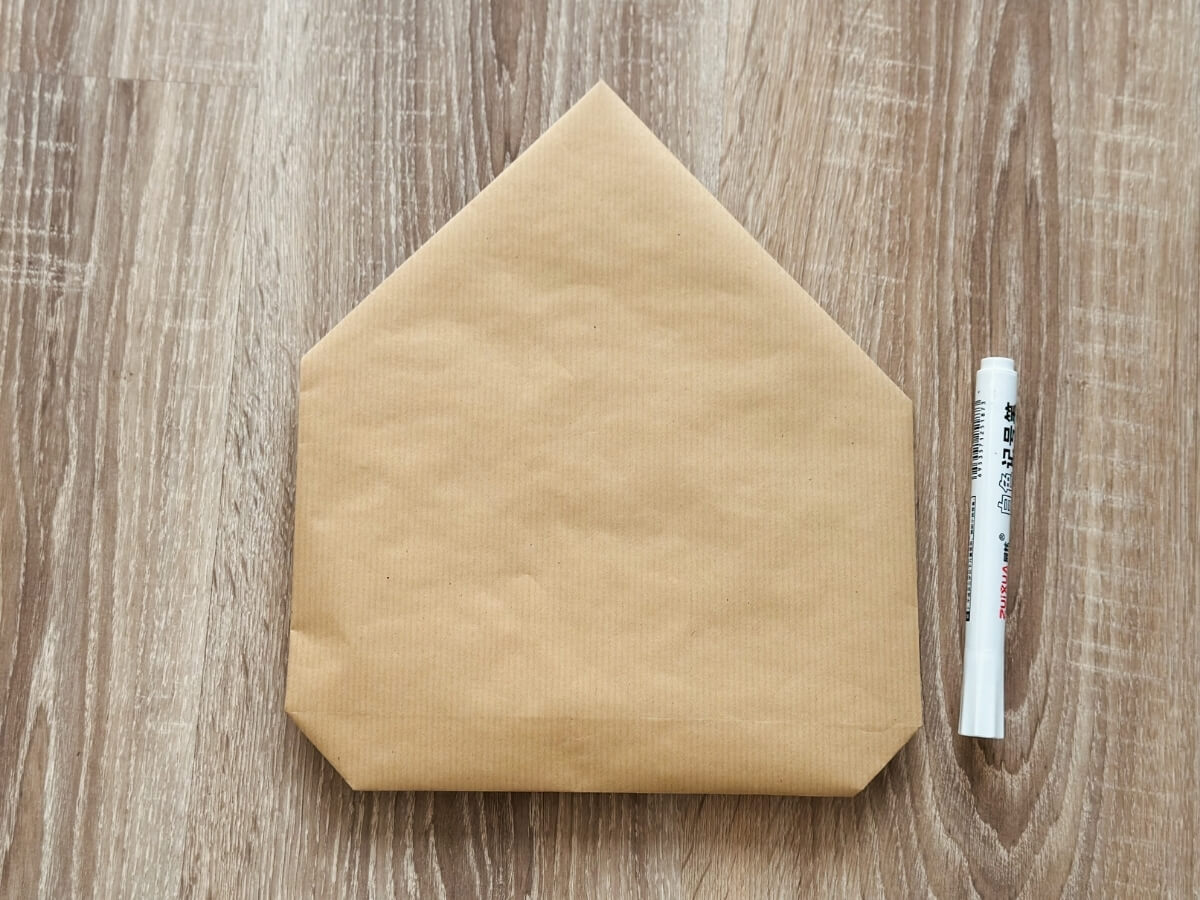 Biela kriedová fixka položená vedľa hnedého baliaceho papiera zloženého do tvaru domčeka.