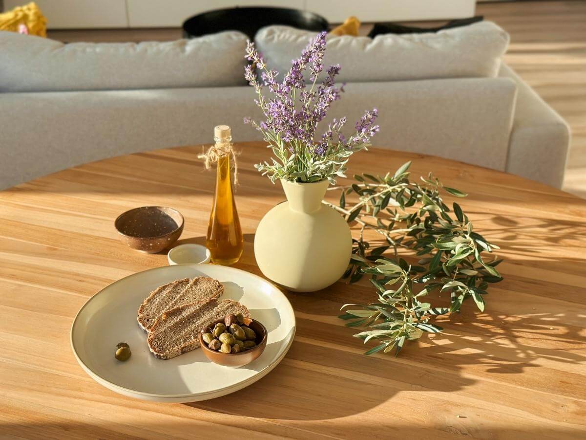 Chlieb s olivami a olivovým olejom vedľa keramickej vázy s levanduľou a konárom olivovníka.