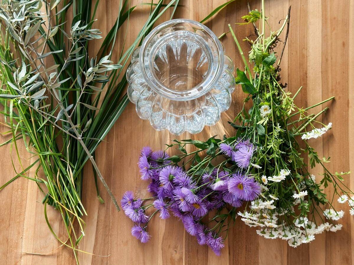 Veľká sklenená váza, kvety a doplnková zeleň položené na stole.