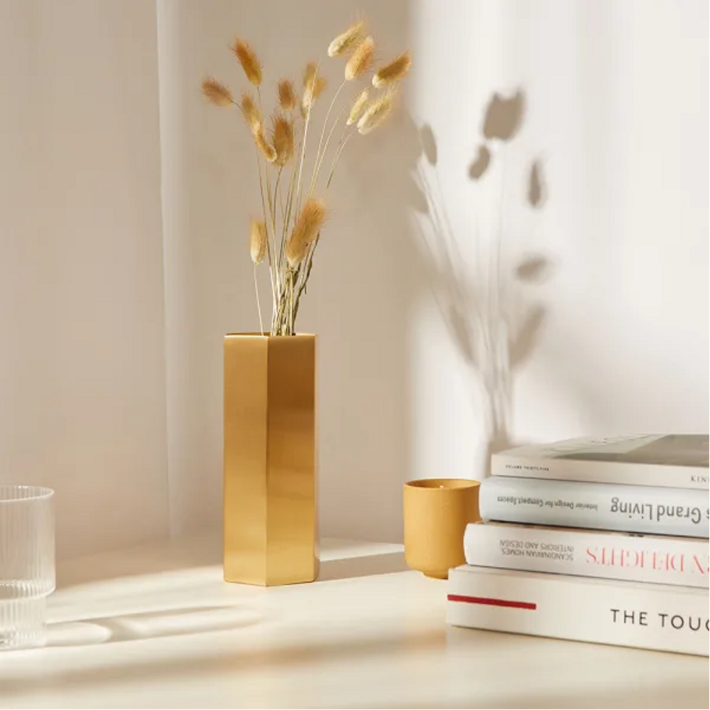 Mosadzná váza so sušenými kvetmi v šesťuholníkovom tvare pri knihách o dizajne