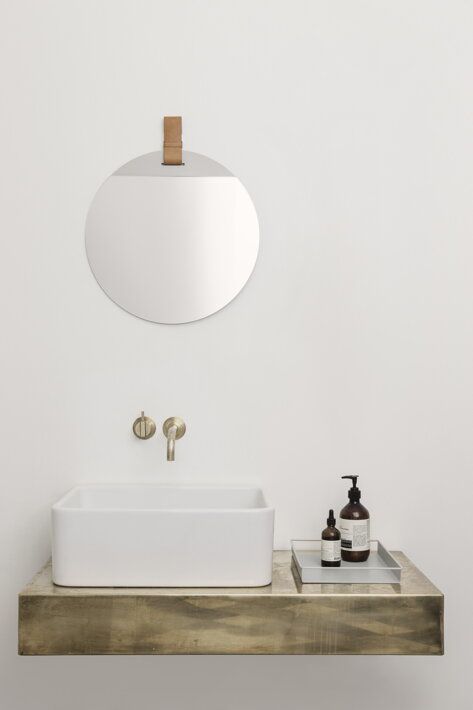 Malé okrúhle nástenné zrkadlo s hnedým koženým remienkom nad kúpeľňovým umývadlom