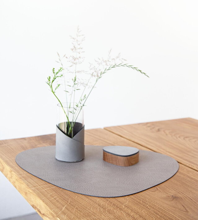 Veľké kožené prestieranie v teplej sivej farbe na drevenom stole s dizajnovou vázou a drevenou soľničkou