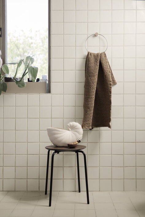 Minimalistický okrúhly chrómový vešiak s hnedým uterákom v béžovej kúpeľni pri dizajnovej váze