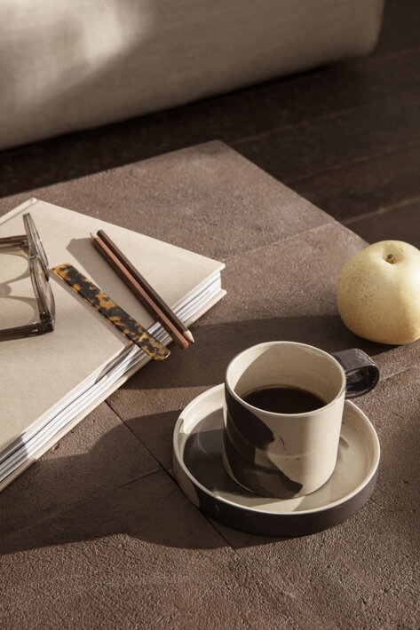 Dvojfarebná šálka s kávou na veľkej podšálke na stole s knihou