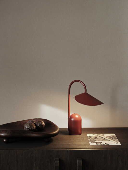 Prenosná lampa v hrdzavo červenej farbe so zapnutým svetlom na komode