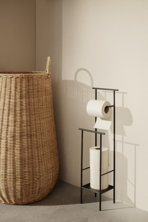 Ratanový kôš postavený v rohu kúpeľne vedľa stojana s toaletným papierom