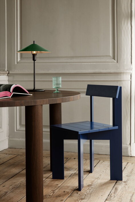 V miestnosti s drevenou podlahou je masívny okrúhly jedálenský stôl, na ňom položená stolová lampa, kniha a pohár vody a prisunutá modrá drevená stolička