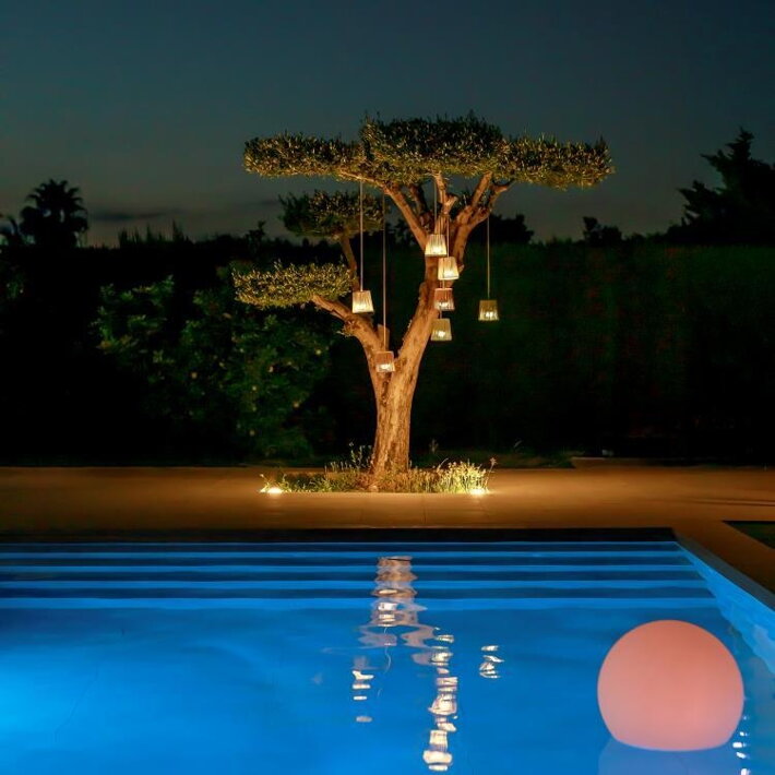 Viacero rozsvietených mini závesných lámp na strome pred bazénom vo večernej atmosfére