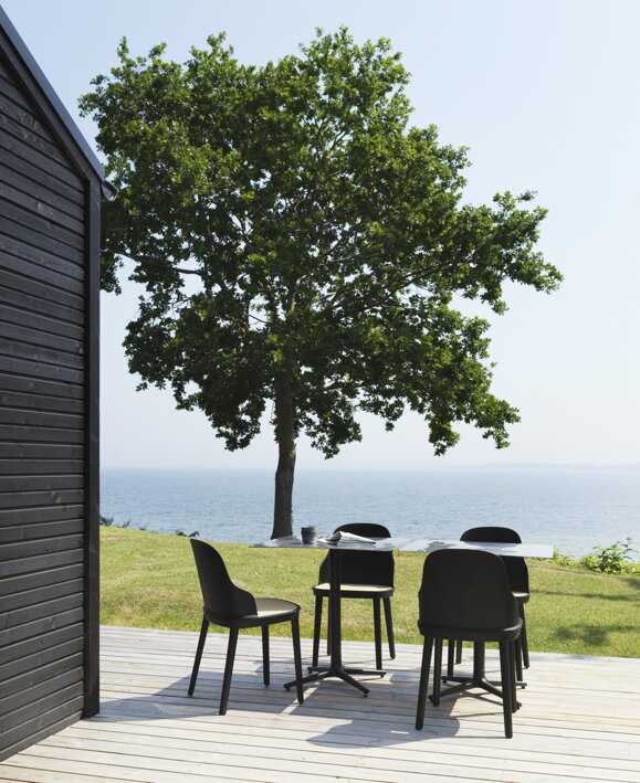 Dva štvorcové kaviarenské stoly so 4 stoličkami na drevenej terase neďaleko morského pobrežia so zeleňou a morom v pozadí