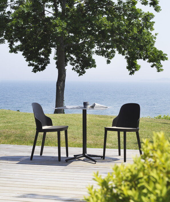 Štvorcový kaviarenský stôl s dvoma stoličkami na drevenej terase neďaleko morského pobrežia so zeleňou a morom v pozadí