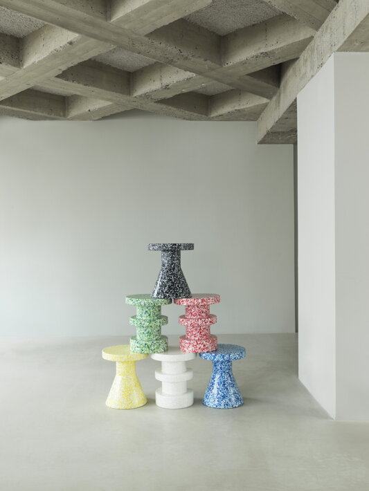 Sedem príručných stolíkov v rôznych farbách postavených na sebe v industriálnom priestore
