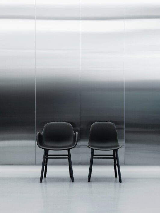 Dve čierne stoličky s plastovým sedákom a podrúčkami a čiernymi dubovými nohami umiestnené pred veľkou chromovanou stenou