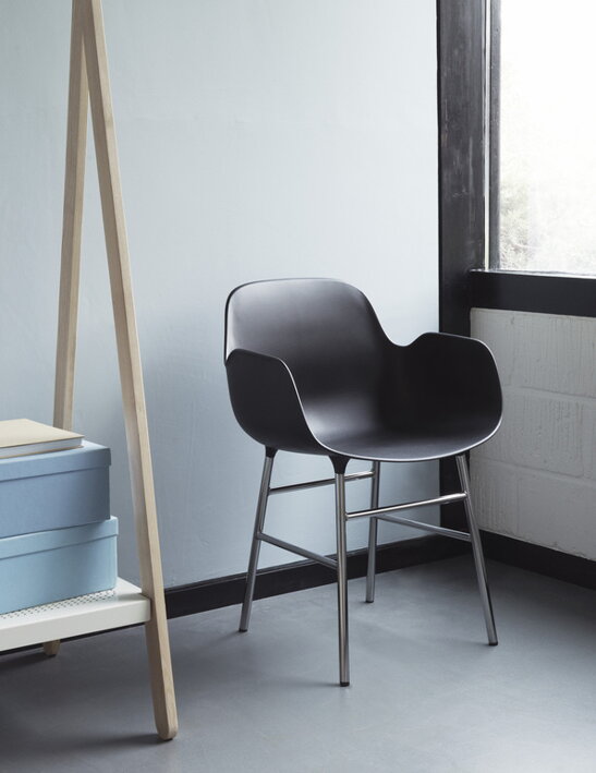 Čierna plastová stolička s podrúčkami a s nohami z chrómu umiestnená vedľa dreveného regálu a okna