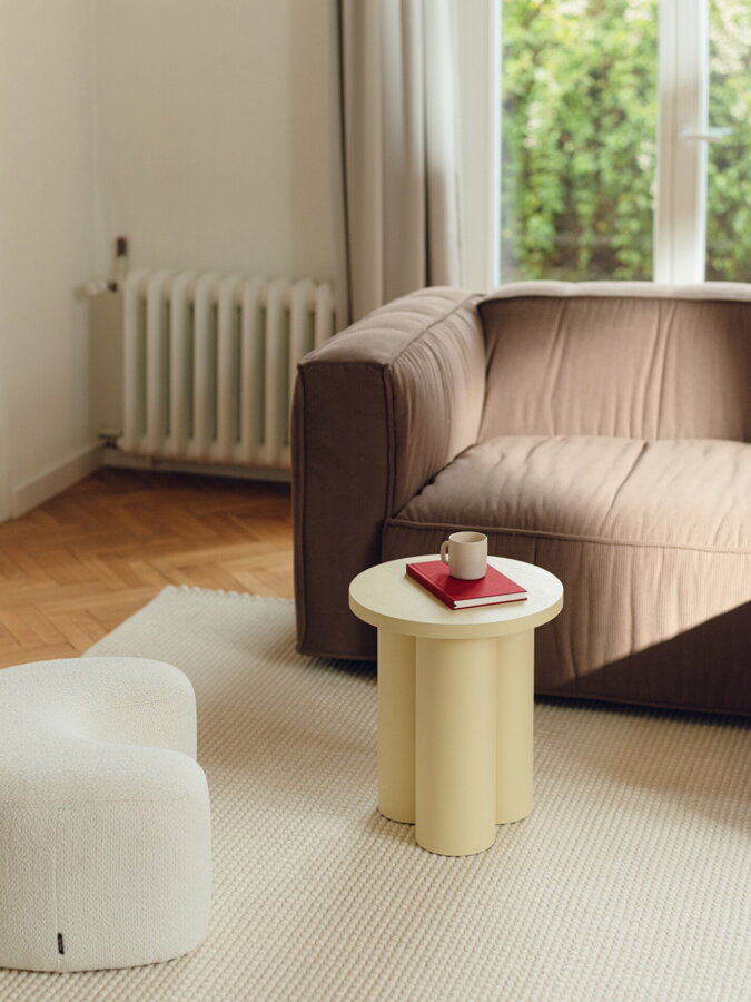 Svetložltý príručný stolík, na ktorom je položená kniha a biela šálka, v obývačke vedľa hnedej sedačky a bieleho pufu.