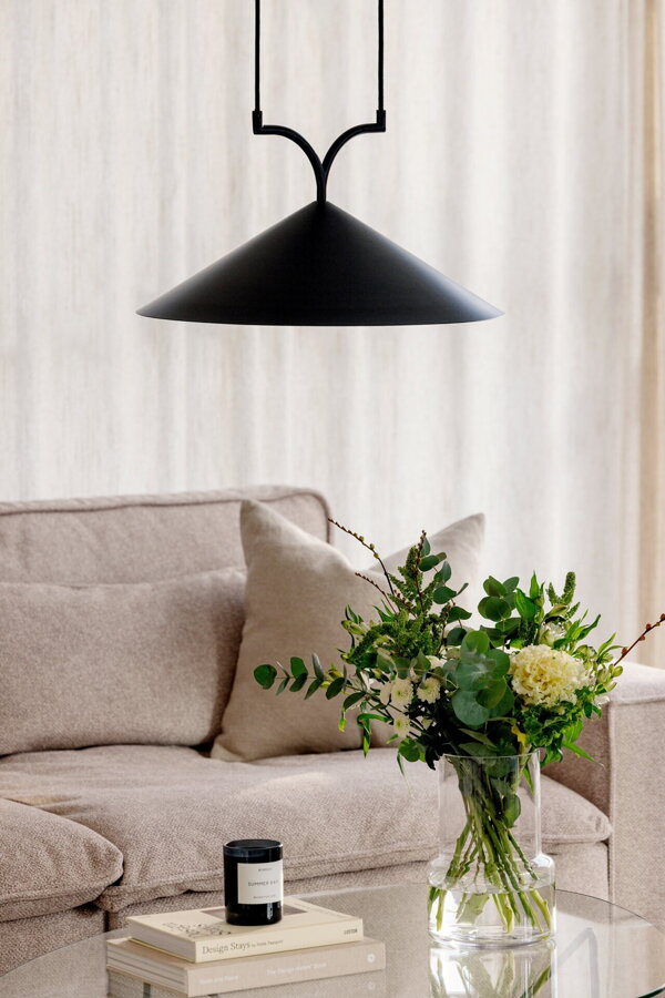 Čierna závesná lampa s trojuholníkovým tienidlom nad preskleným konferenčným stolíkom, na ktorom je váza s kyticou kvetou.