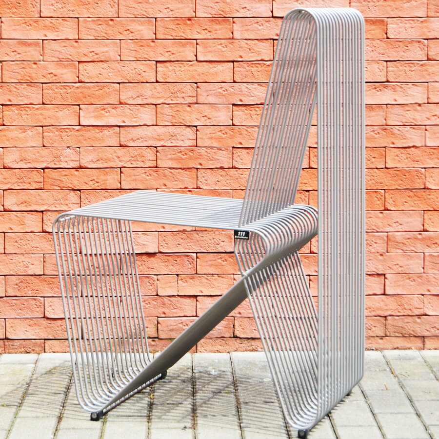 Dizajnová kovová záhradná stolička XY striebornej farby pred tehlovou stenou.