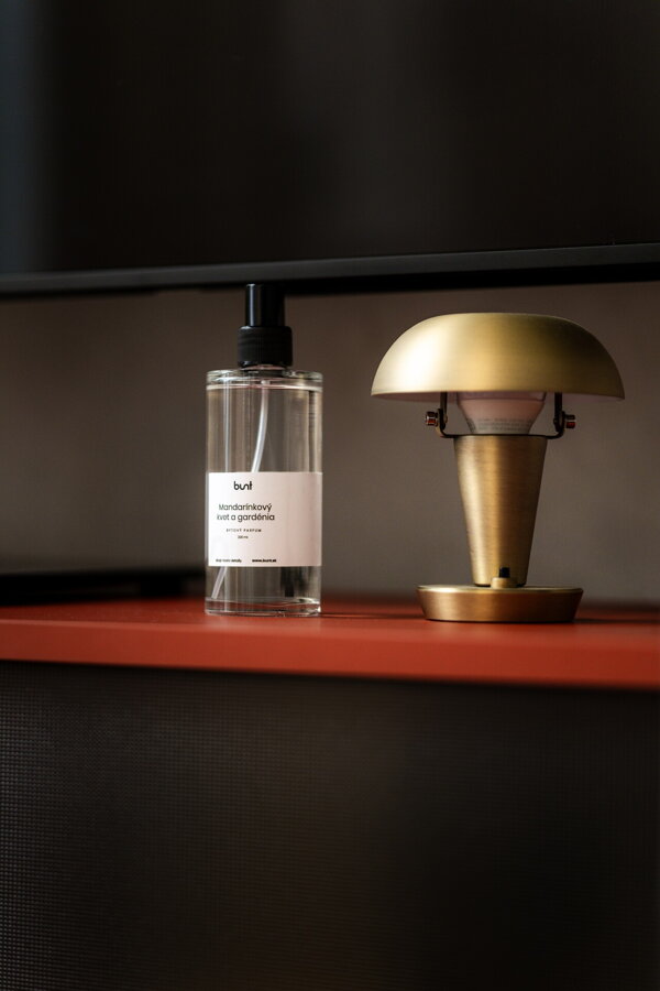 Bytový parfum s vôňou mandarínkového kvetu a gardénie v čírej sklenenej fľaštičke na červenej komode vedľa mosadznej stolovej lampy.