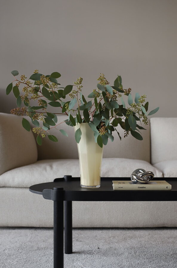 Kytica kvetov v maslovo žltej sklenenej váze s hexagonálnou základňou na tmavohnedom konferenčnom stolíku pred bielou sedačkou.