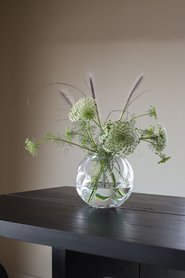 Okrúhla číra sklenená váza s kyticou kvetou na čiernom stolíku.