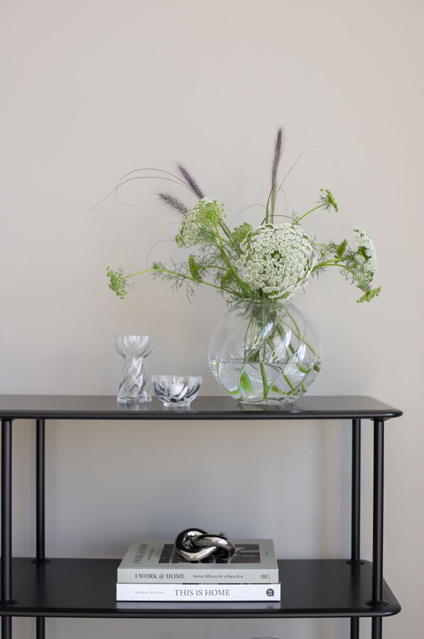 Okrúhla číra sklenená váza s kyticou kvetov spolu s vázou a miskou z rovnakej kolekcie, na čiernom stolíku.s poličkou.