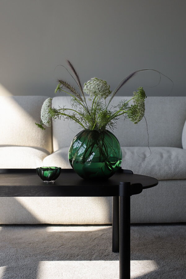 Okrúhla smaragdovozelená sklenená váza s kyticou bielych kvetov na tmavohnedom konferenčnom stolíku pred bielou sedačkou.