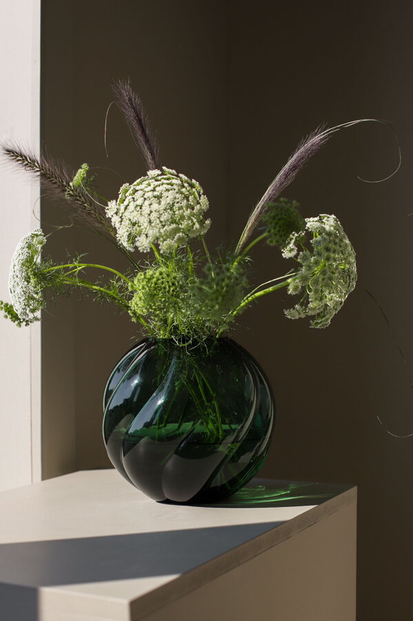 Okrúhla smaragdovozelená sklenená váza s kyticou bielych kvetov na bielom podstavci v žiare slnečných lúčov.