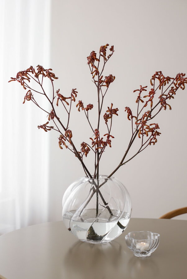 Číra sklenená váza s červenými kvietkami a vedľa nej číry svietnik na čajovú sviečku.