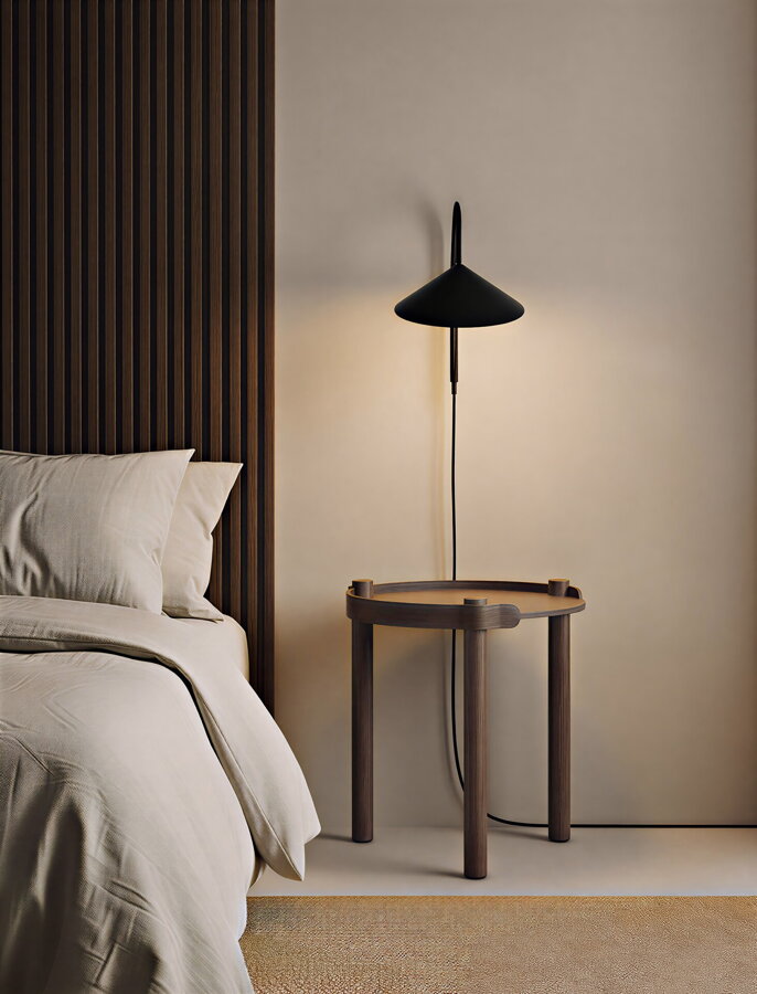 Okrúhly príručný stolík Woody Table z tmavohnedého dubového dreva pod nočnom lampou vedľa postele v spálni.