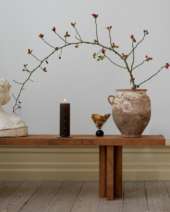 Hnedá adventná valcová sviečka na konferenčnom stolíku s hlinenou vázou s halúzkou.