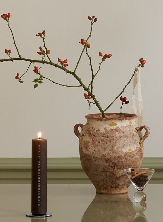 Horiaca hnedá adventná sviečka na stole vedľa pohára s červeným vínom a hnedej hlinenej vázy s vetvičkou.