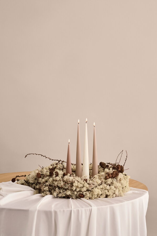 Štyri kónické sviečky hnedej a bielej farby v béžovom dekoračnom venci, položenom na okrúhlom stole s dlhým bielym obrusom.