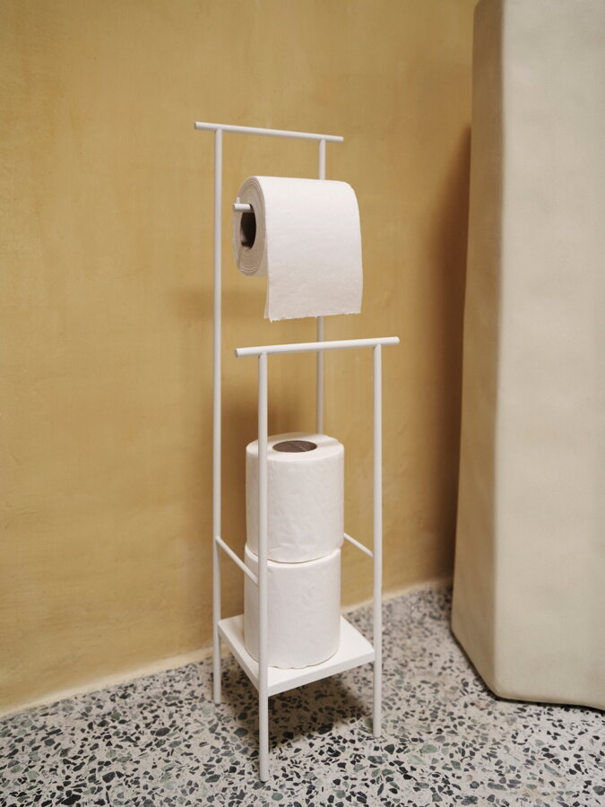 Biely oceľový stojan na toaletný papier s 3 rolkami papiera, postavený na podlahe vedľa žltej steny