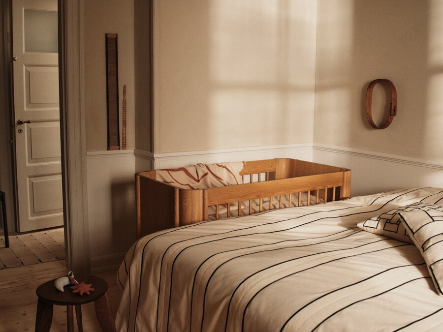 Hnedý príručný stolík z masívneho orecha v spálni vedľa manželskej postele a drevenej detskej postielky.