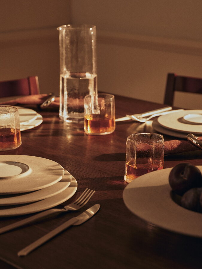 Sklenený pohár a karafa na prestretom stole vedľa kameninových tanierov.