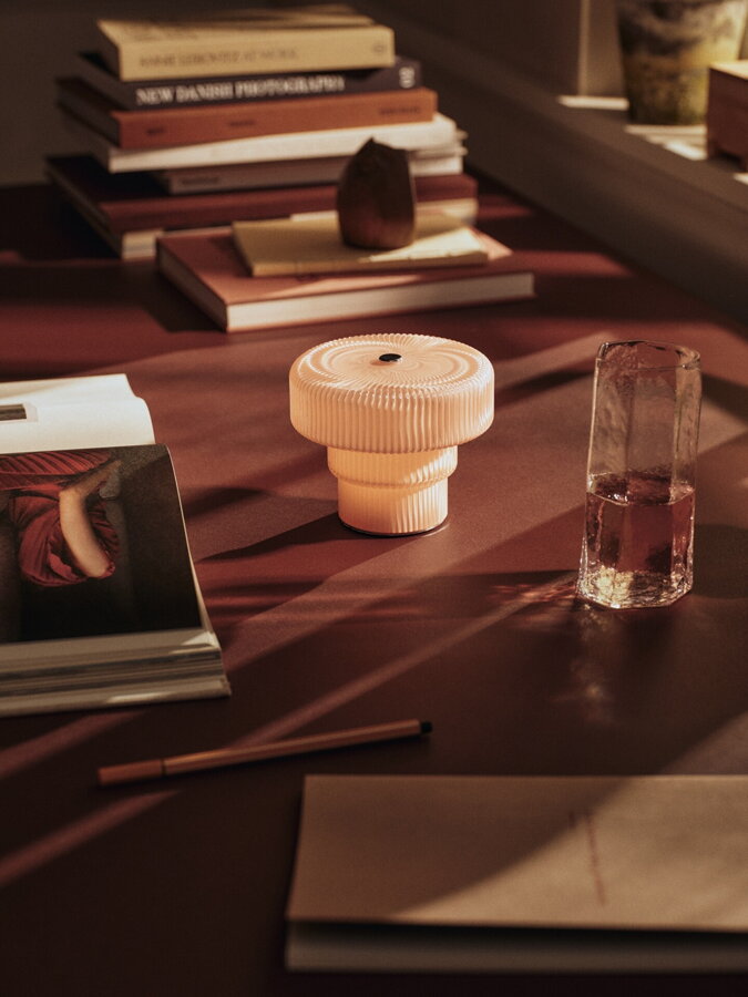 Svetloružová prenosná lampa na stole vedľa kôpky kníh a pohára s nápojom.