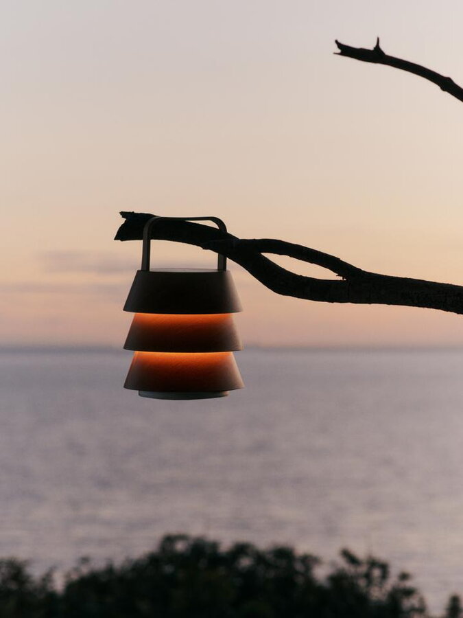 Prenosná lampa s dreveným tienidlom zavesená na konári v pozadí s morom.