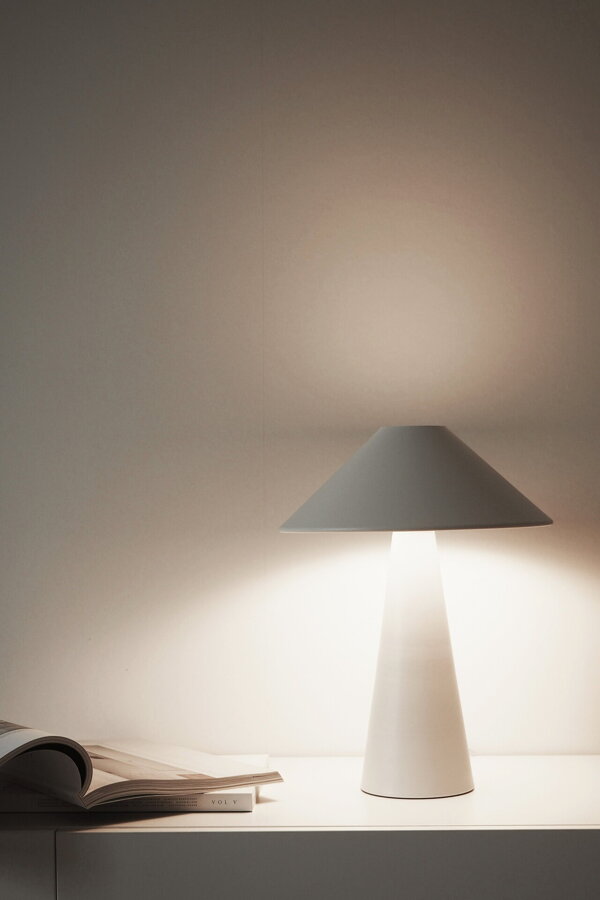 Svietiaca biela kovová stolová lampa na bielej skrinke vedľa otvorenej knihy.