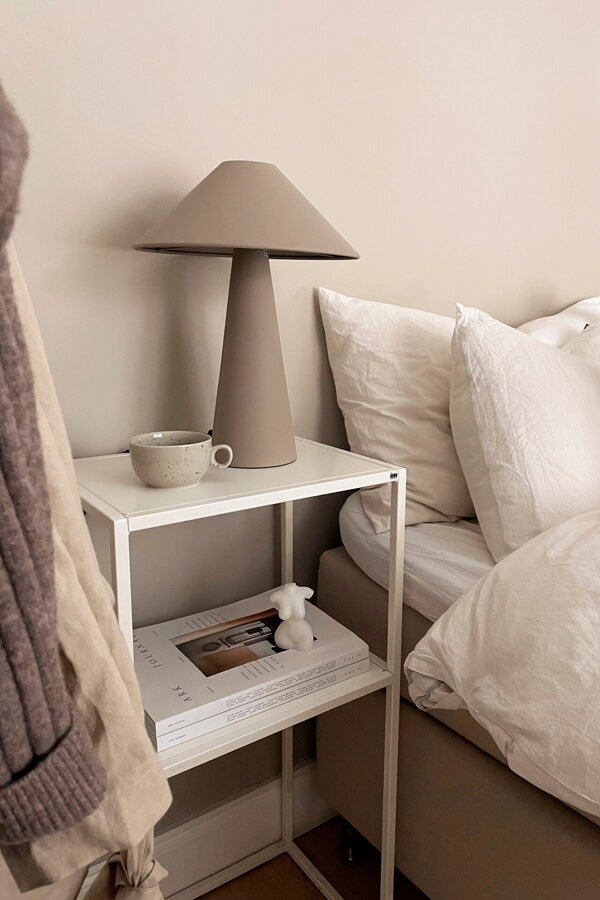 Svetlá hnedosivá stolová lampa Cannes z matného lakovaného kovu na bielom príručnomm stolíku vedľa postele.