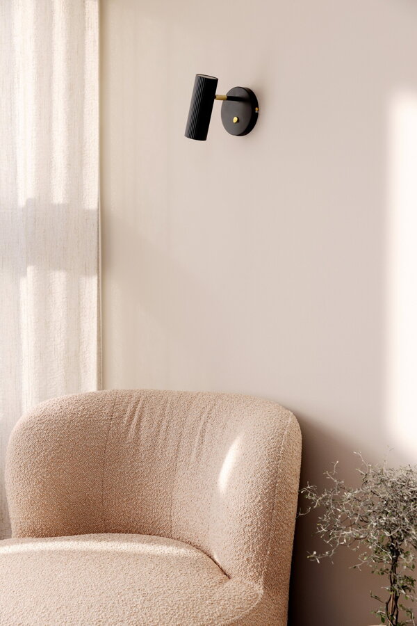 Čierna nástenná lampa na stene nad béžovým čalúneným kreslom.