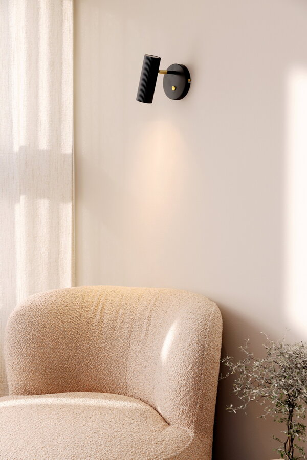 Čierna nástenná lampa so svetlom nasmerovaným na béžové čalúnené kreslo pod lampou.