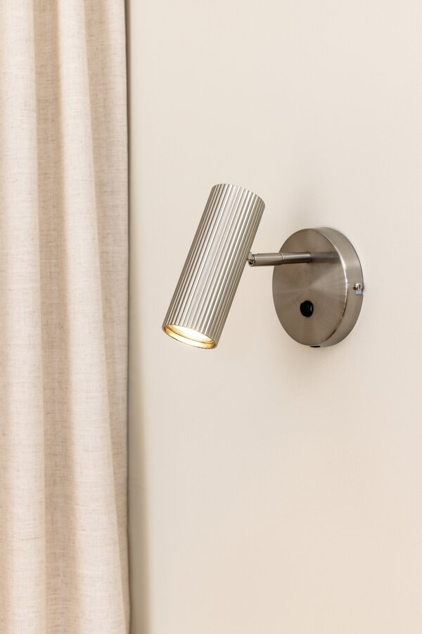 Polohovateľná kovová nástenná lampa Hubble v prevedení brúsenej ocele na béžovej stene.