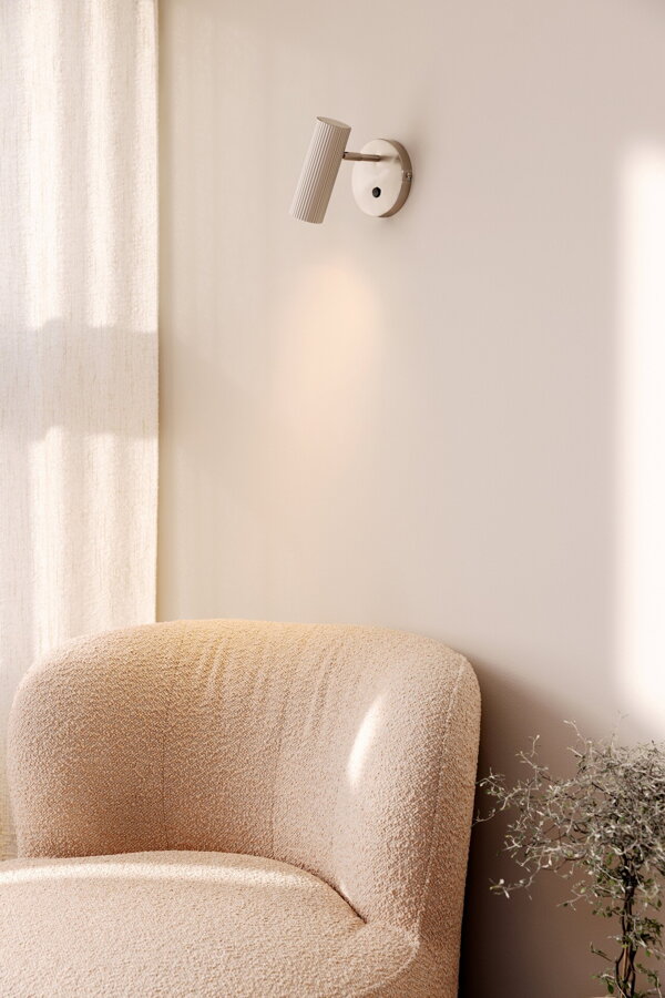 Polohovateľná kovová nástenná lampa Hubble v prevedení brúsenej ocele nad béžovým čalúneným kreslom.