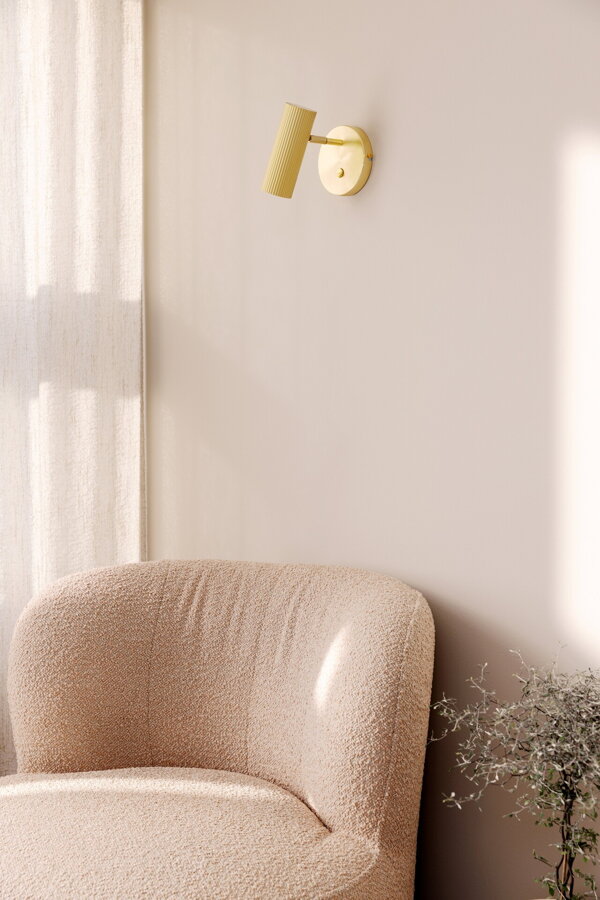 Polohovateľná kovová nástenná lampa Hubble v prevedení brúsenej mosadze nad béžovým čalúneným kreslom.