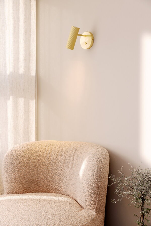 Svietiaca polohovateľná kovová nástenná lampa Hubble v prevedení brúsenej mosadze nad béžovým čalúneným kreslom.