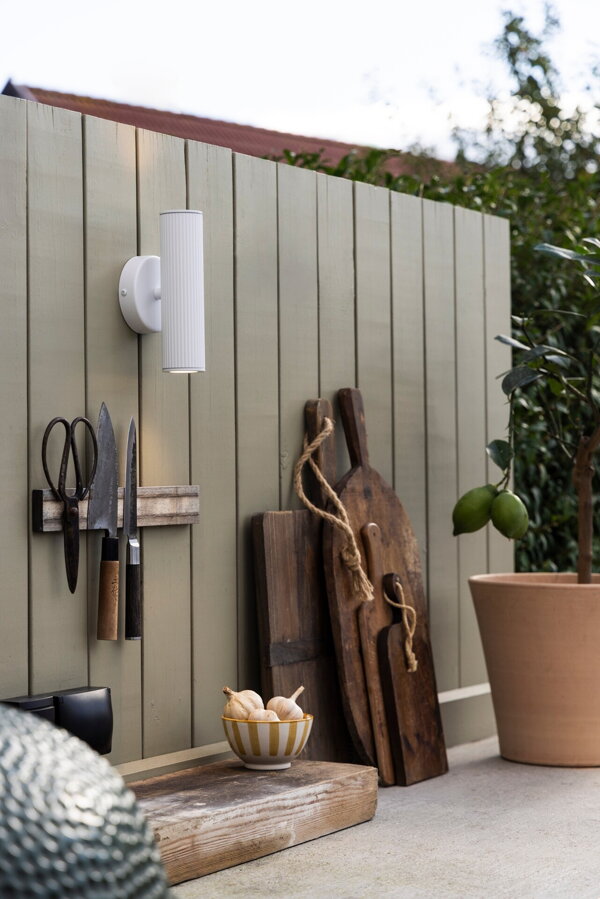 Biela  hliníková nástenná lampa s obojsmerným svietením vo farbe brúsenej ocele, zavesená na drevenej stene na terase.